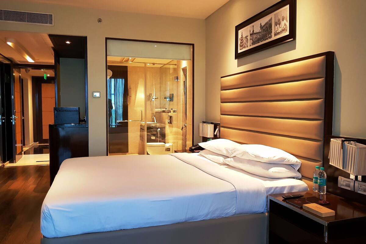 Una habitación de hotel con una cama grande, una mesita de noche y un baño visible a través de un separador de vidrio.