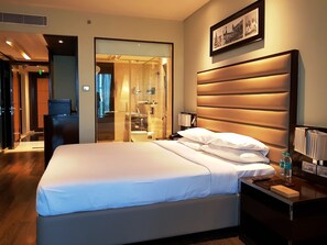 Una habitación de hotel con una cama grande, una mesita de noche y un baño visible a través de un separador de vidrio.