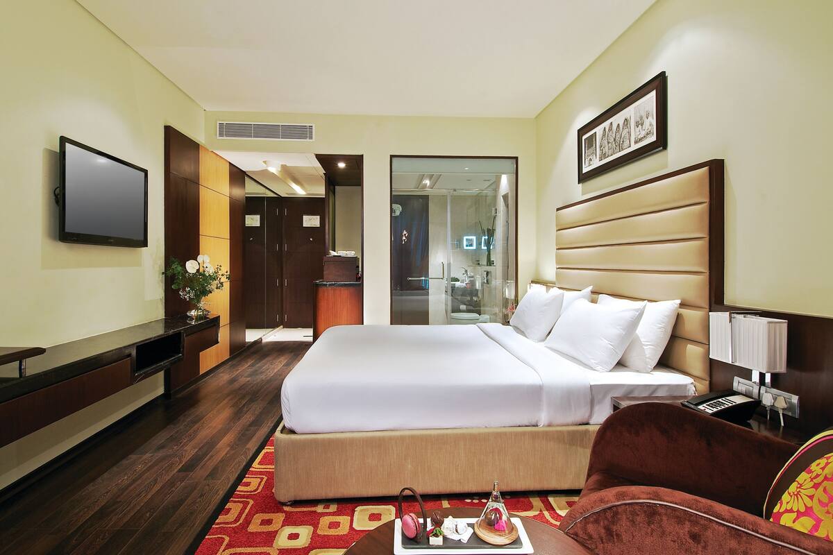 Habitación de hotel con una cama grande, televisor, escritorio con computadora y baño visible a través de una puerta de vidrio.