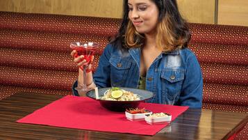 Une personne est assise à une table devant un bol de nourriture, un verre d’une boisson rouge et une petite assiette de condiments.