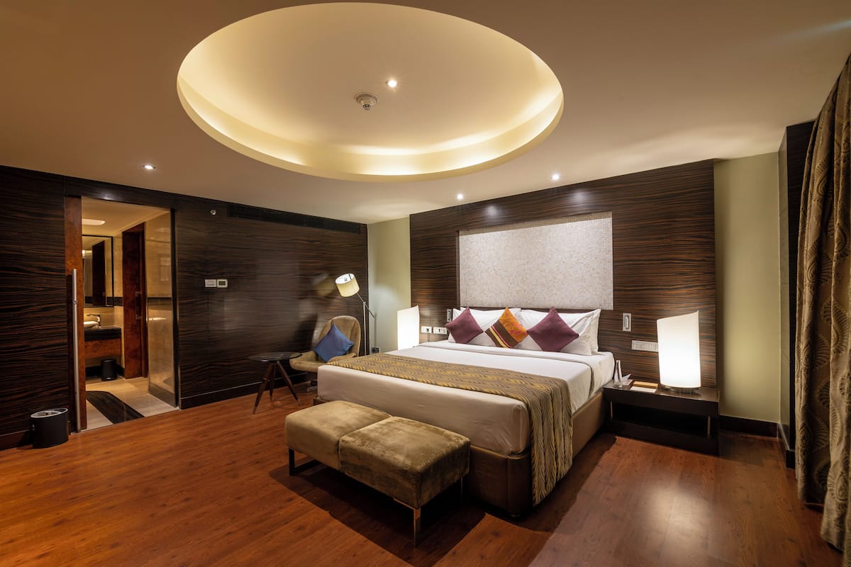 Una habitación de hotel moderna con una cama grande, una mesita de noche con lámpara, un banco y una lámpara de techo.