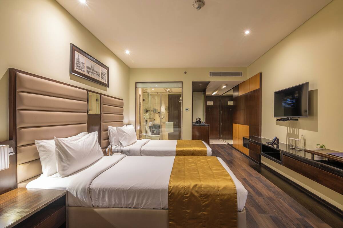 Habitación de hotel con dos camas, un televisor, un escritorio y un baño visible a través de una puerta de cristal.