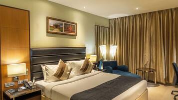 A hotel room with a large bed, a bedside table with a lamp, a chair, a desk, and a curtain.