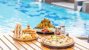Una mesa de comedor con un sándwich, papas fritas, ensalada y pizza junto a una piscina.