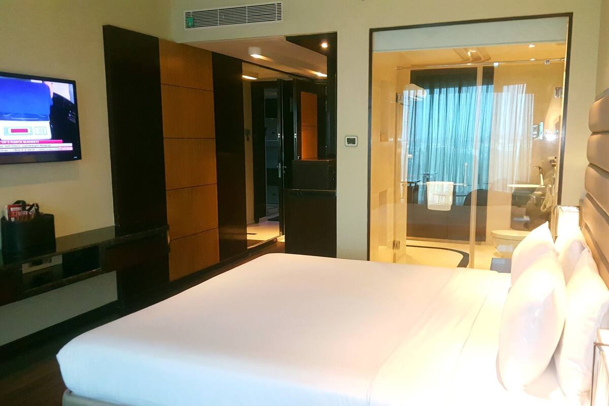 Habitación de hotel con una cama grande, televisor montado en la pared y baño visible a través de un vidrio.
