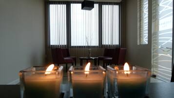 Una habitación con tres velas encendidas sobre una mesa, dos sillas y una ventana con persianas verticales.