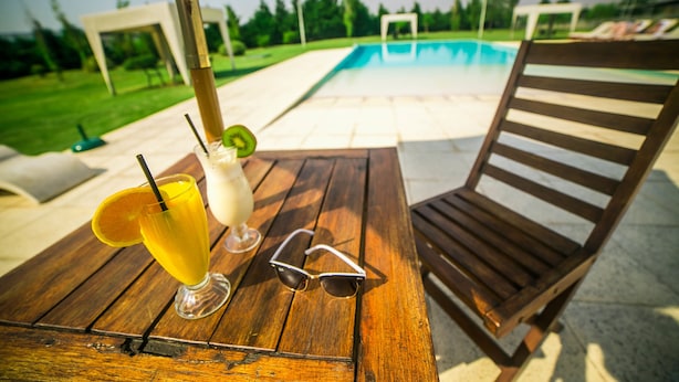 A wooden table with a drink, sunglasses, and a pool in the background.