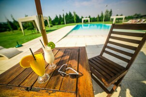 Una mesa de madera con una bebida, gafas de sol y una piscina al fondo.