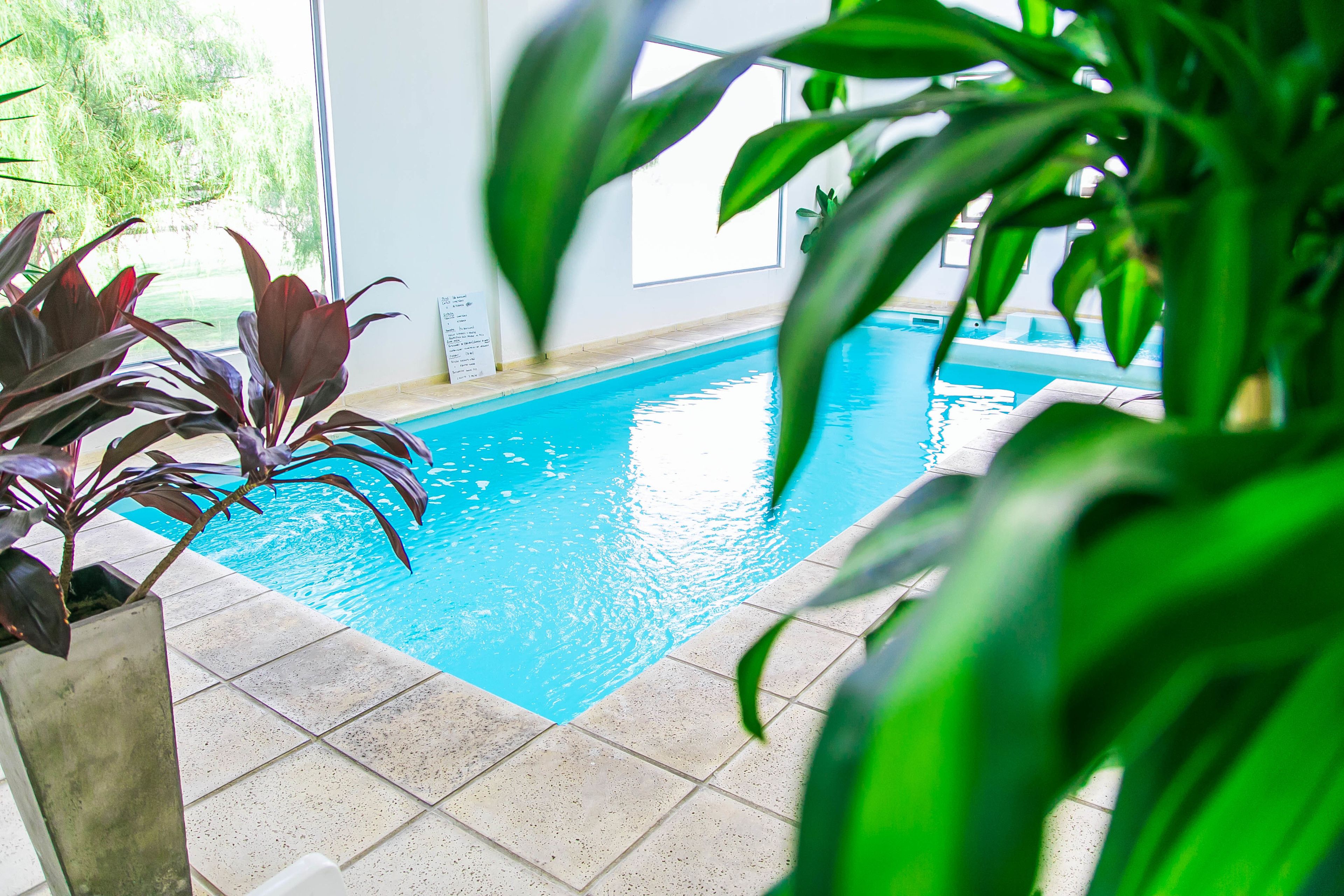 An indoor swimming pool with clear blue water, surrounded by tiled flooring and potted plants.
