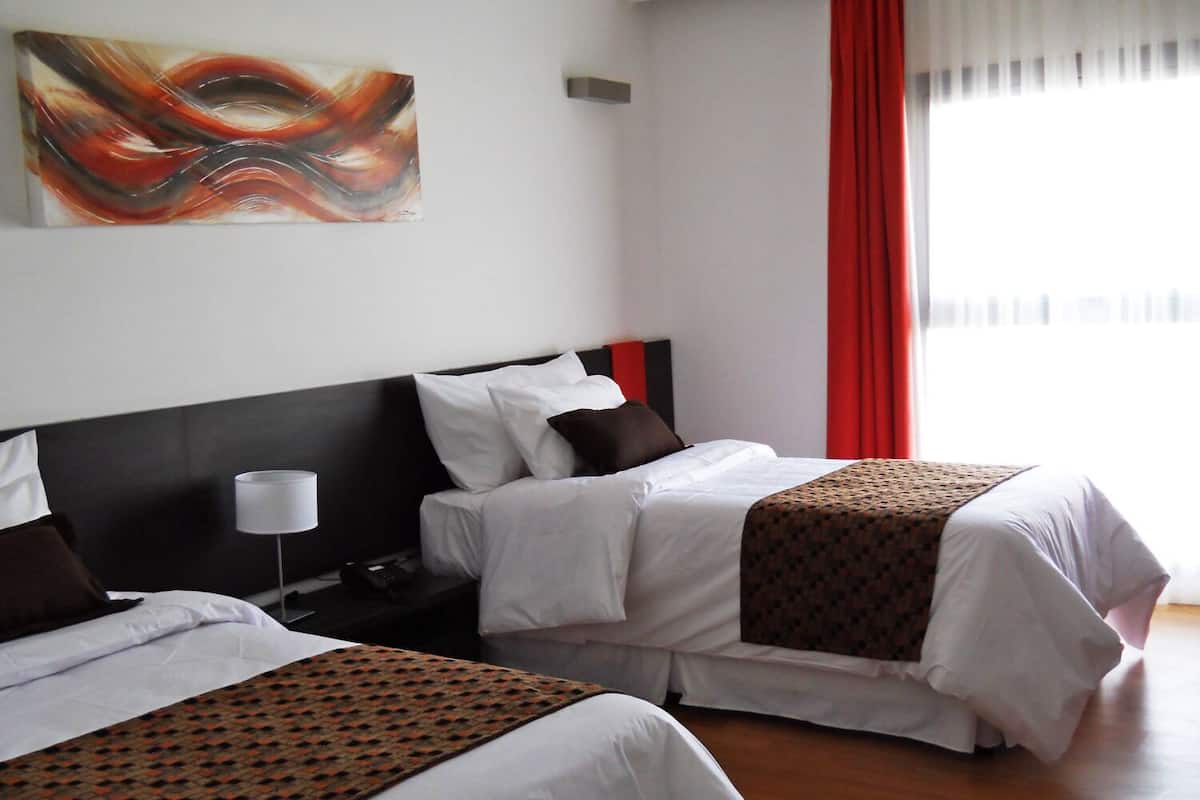 Habitación de hotel con dos camas, una mesita de noche con lámpara, un cuadro en la pared y una ventana con cortinas rojas.