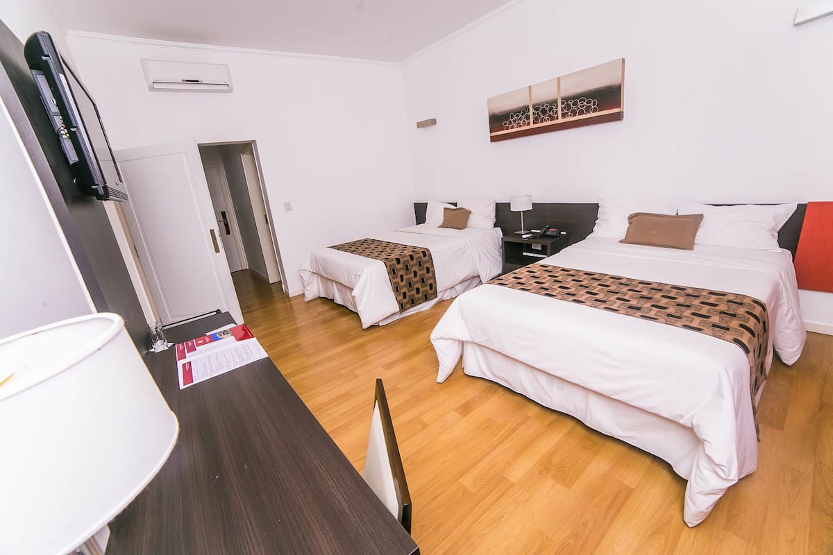 Habitación de hotel con dos camas, un escritorio, una silla, una lámpara y una puerta que da a otra habitación.