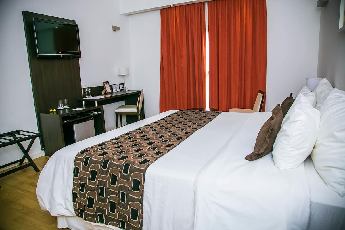 Habitación de hotel con cama, escritorio, televisor y ventana con cortinas rojas.