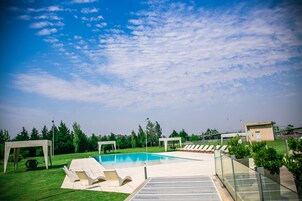 Piscina con sillas de descanso, un área de estar techada y un pequeño edificio en un entorno exterior ajardinado.