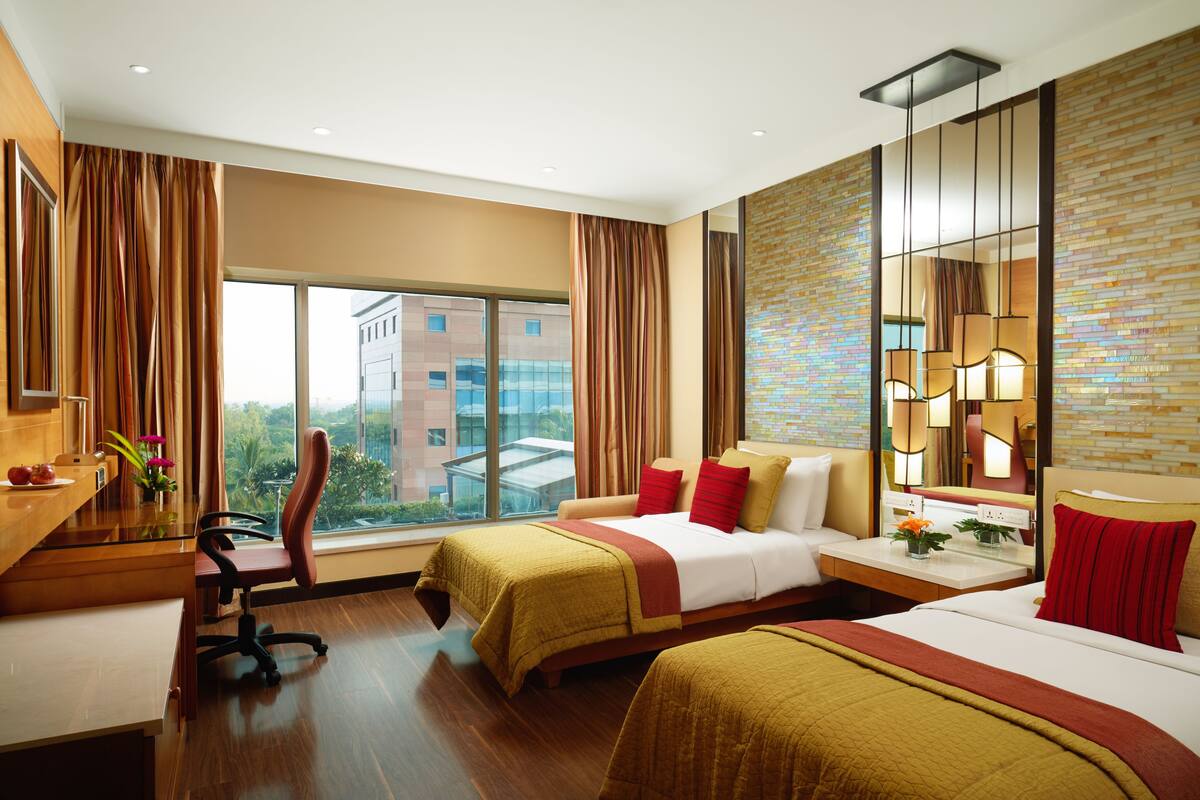 Quarto de hotel com duas camas, uma escrivaninha, uma cadeira e uma grande janela com vista para a cidade.