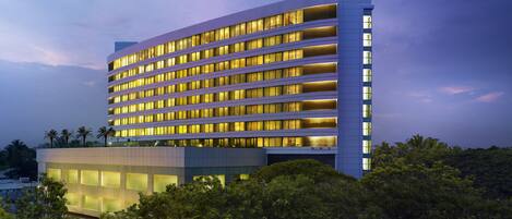 A modern hotel with multiple floors and a flat roof, surrounded by dense greenery.