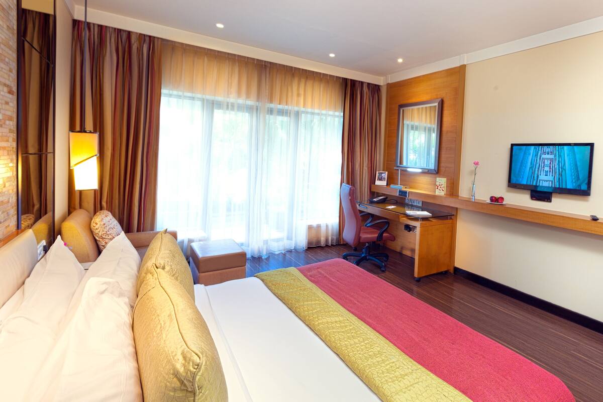 Quarto de hotel com uma cama grande, uma escrivaninha, uma cadeira, televisão e janela com cortinas.