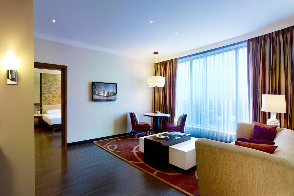 Quarto de hotel com cama, escrivaninha, cadeira, sofá, televisão e uma janela grande com cortinas.