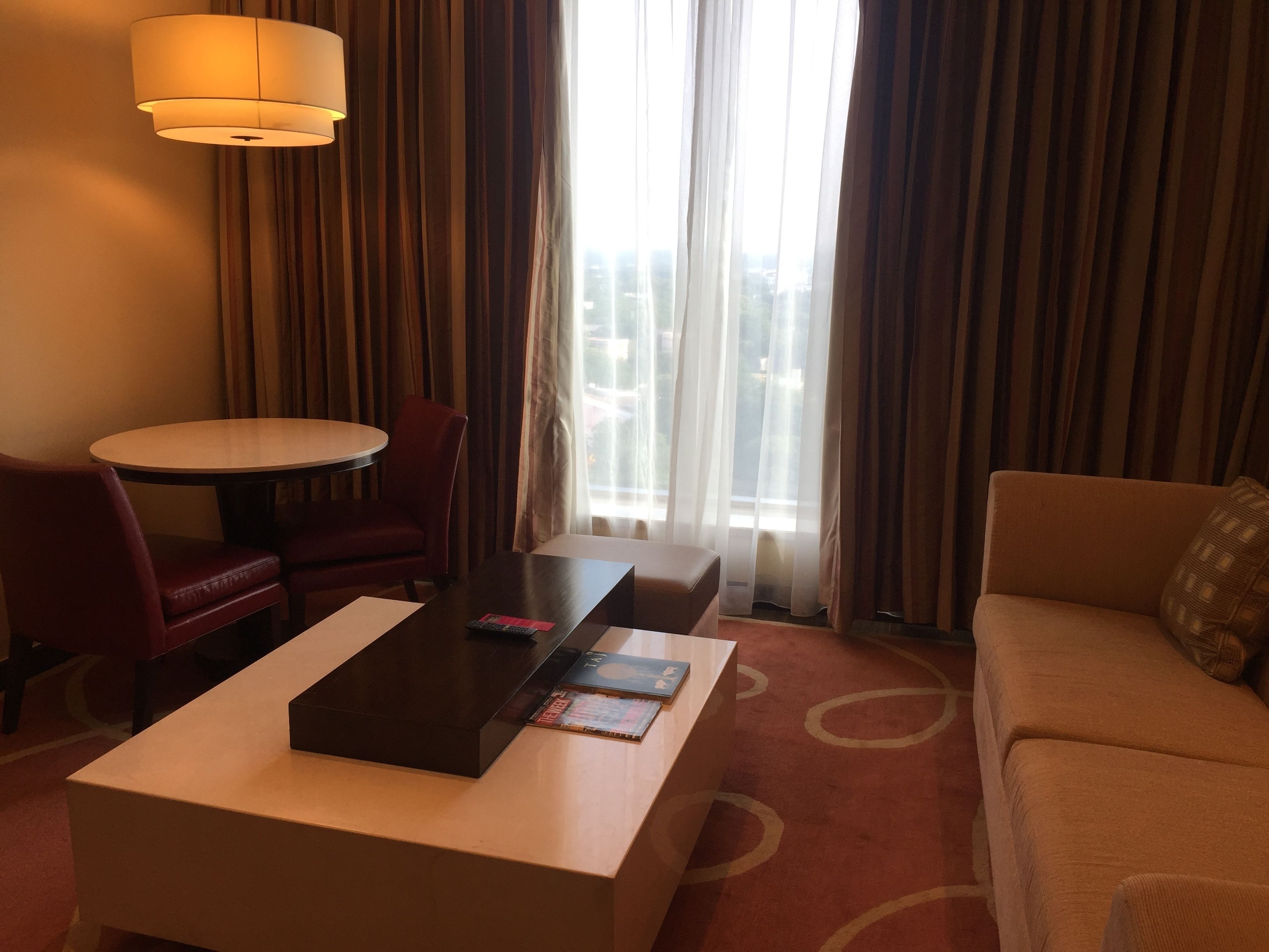 Een hotelkamer met een ronde eettafel, een bank, een salontafel en uitzicht op de stad door grote ramen met gordijnen.