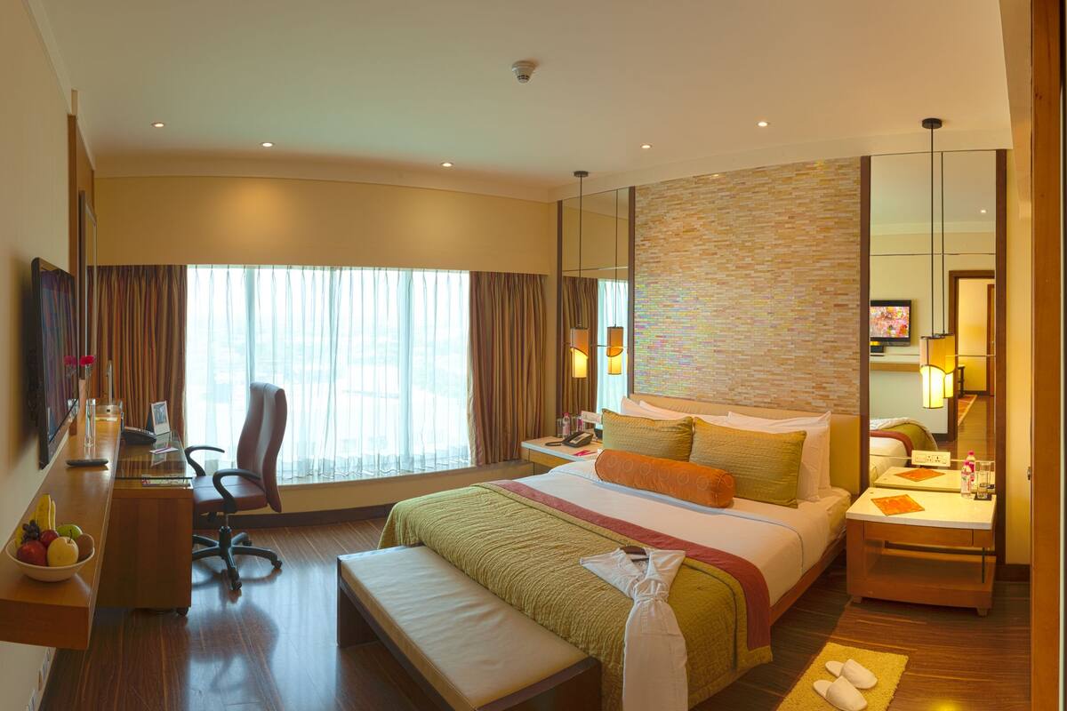 Quarto de hotel com cama, escrivaninha, cadeira e uma janela grande.