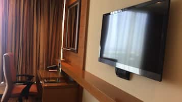 Habitación de hotel con televisión de pantalla plana en la pared, un escritorio de madera, una silla moderna y un amplio ventanal con cortinas.