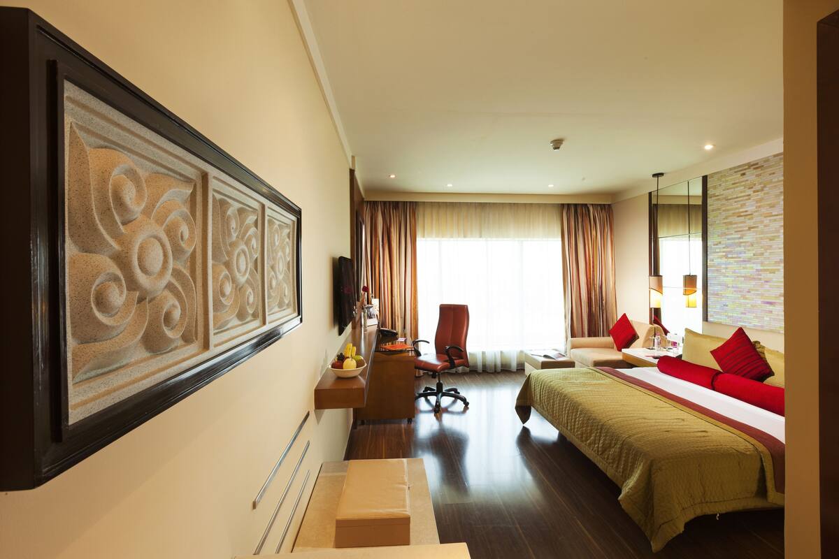 Quarto de hotel com cama, escrivaninha, cadeira, televisão e um grande painel decorativo na parede.