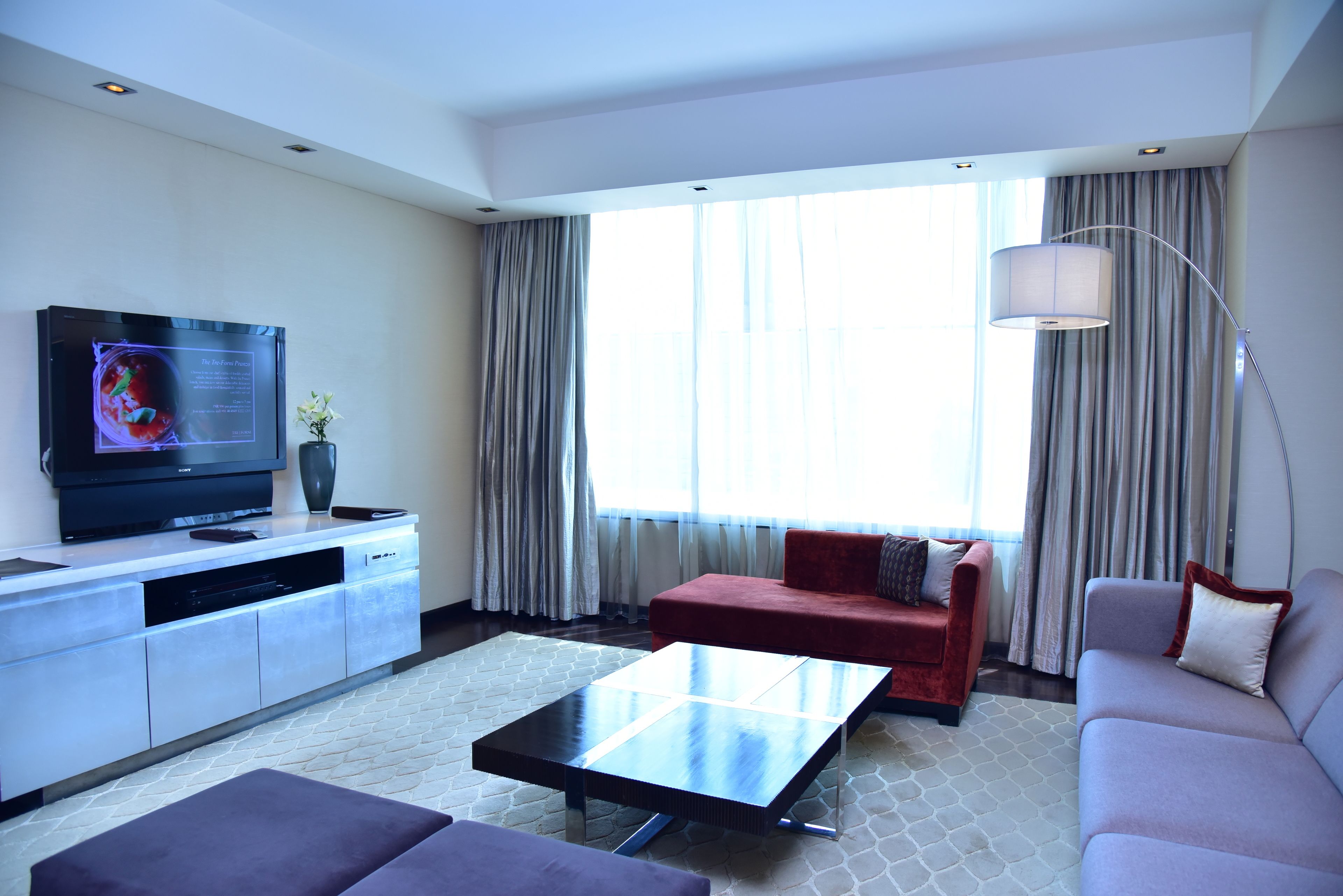 A modern hotel room with a flat-screen TV, a sofa, a red armchair, a coffee table, and a large window with curtains.