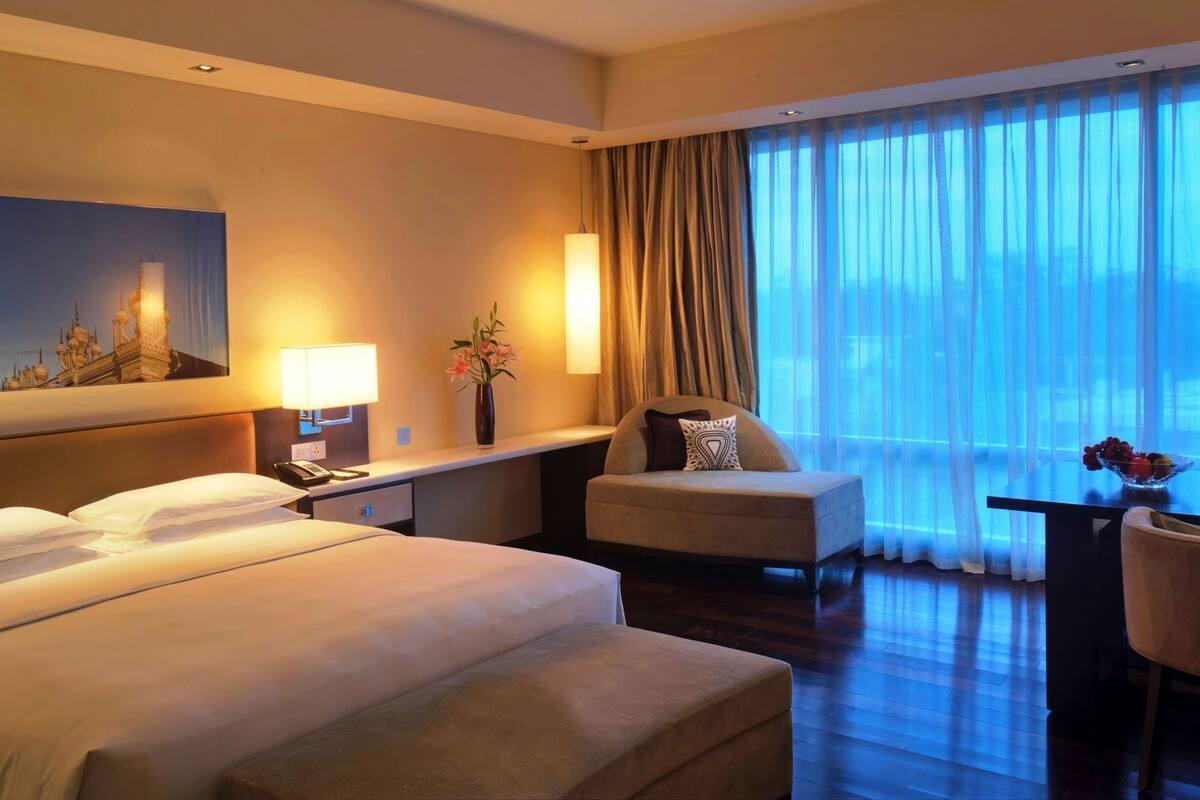 Une chambre d’hôtel avec un lit, un bureau, une chaise, une lampe, un vase de fleurs et un tableau au mur.
