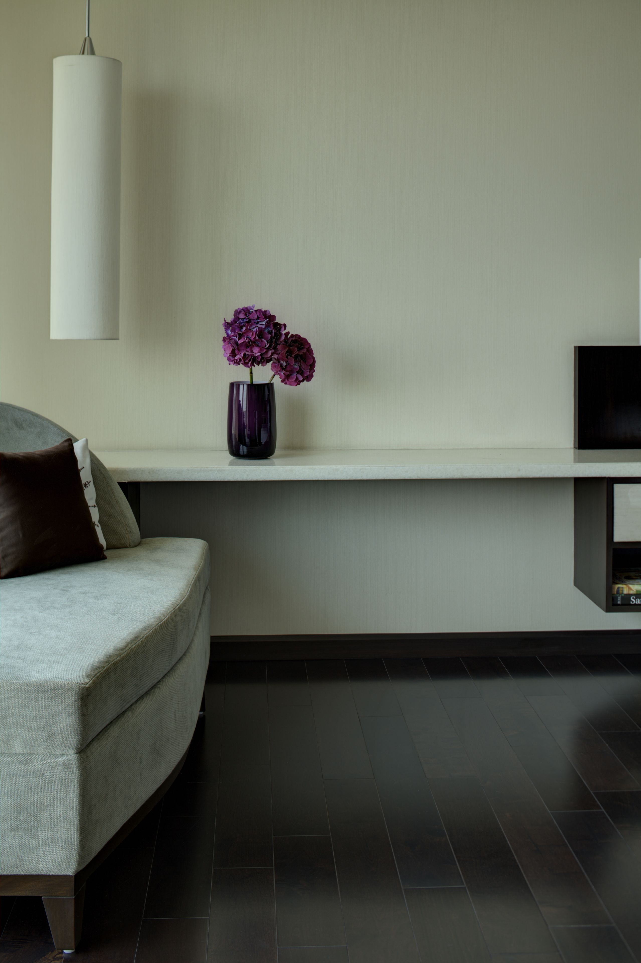 Una sala moderna con un sofá gris, un jarrón con flores moradas, una lámpara colgante blanca y un televisor de pantalla plana sobre una estantería.