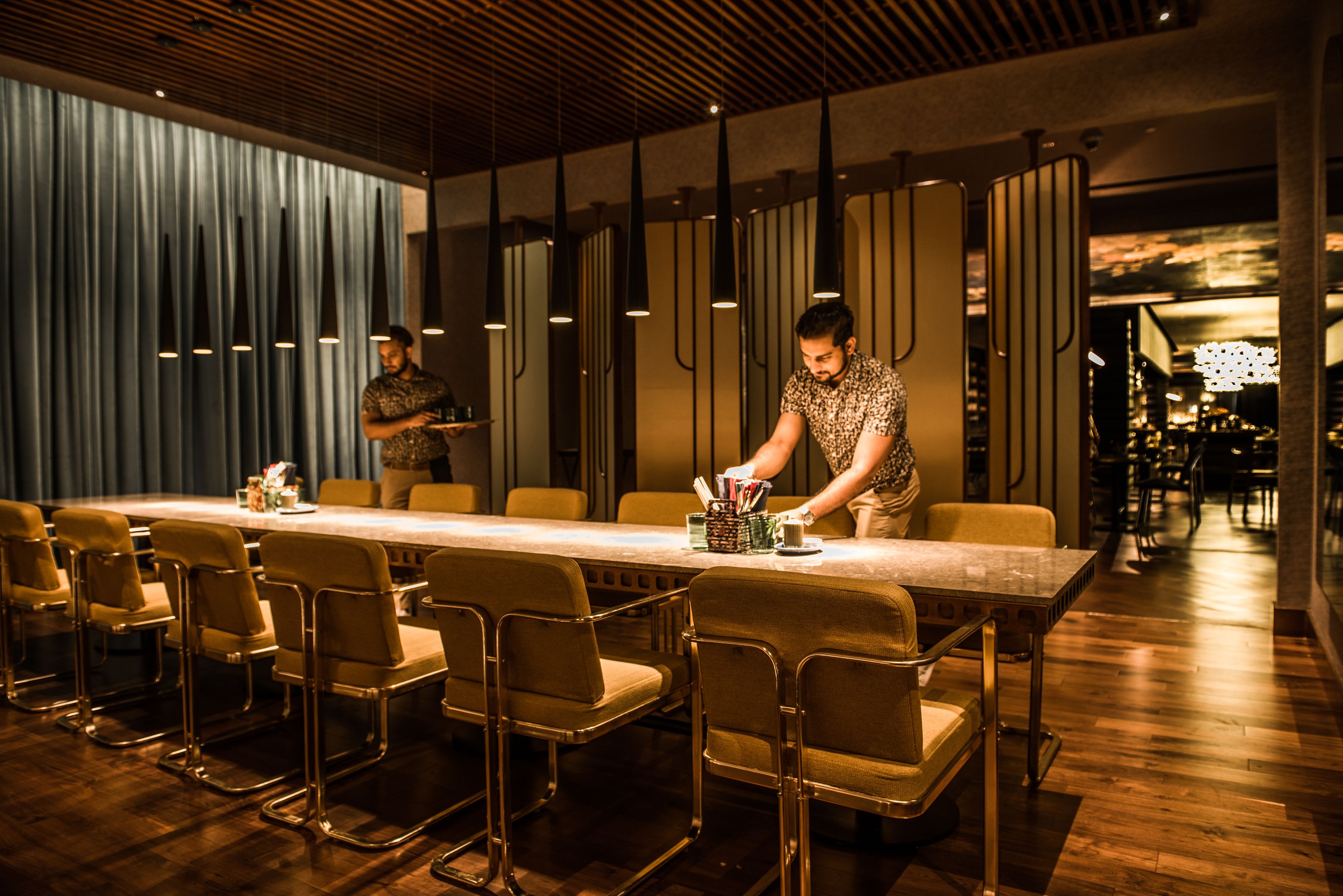 Um restaurante moderno com uma mesa comprida e várias cadeiras, uma pessoa atendendo e um interior com iluminação baixa.