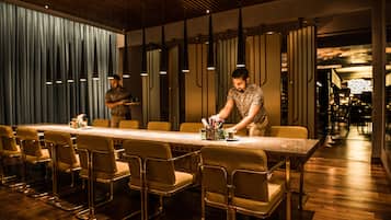 A modern restaurant with a long table and several chairs, a person serving, and a dimly lit interior.