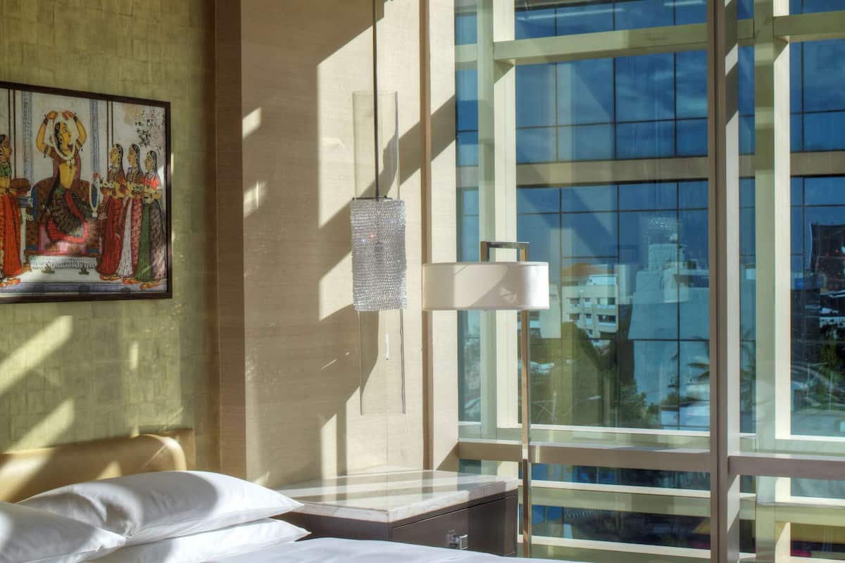 Une chambre d’hôtel avec un grand lit, une fenêtre donnant sur des immeubles, un tableau au mur et une lampe sur une table de chevet.