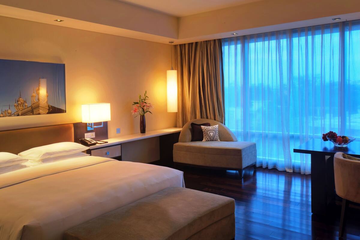 Une chambre d’hôtel avec deux lits, un bureau avec une lampe, un vase de fleurs, une chaise et une vue sur la ville grâce à de grandes fenêtres.