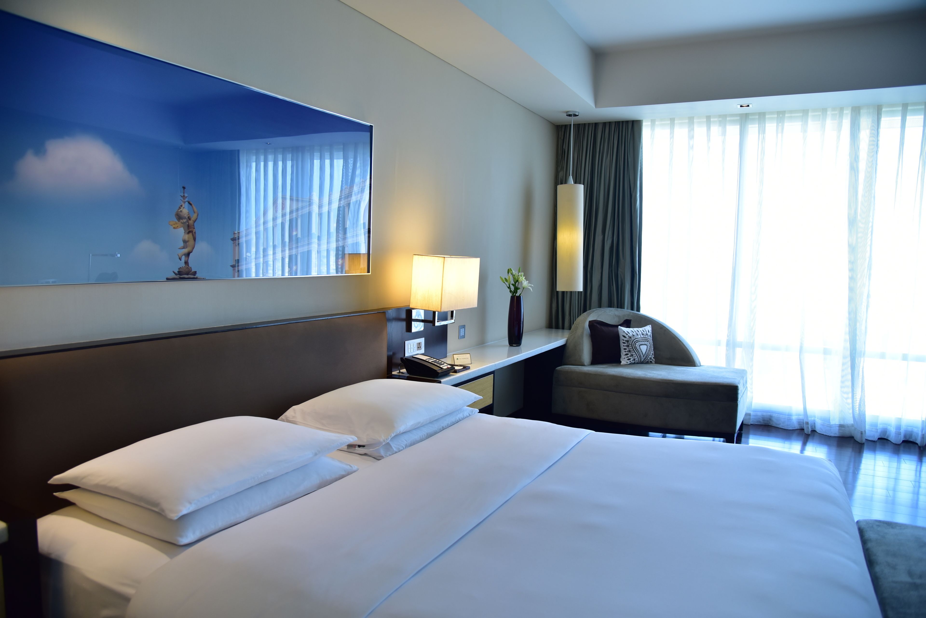 A hotel room with a large bed, a desk with a lamp, a chair, and a large window with curtains.