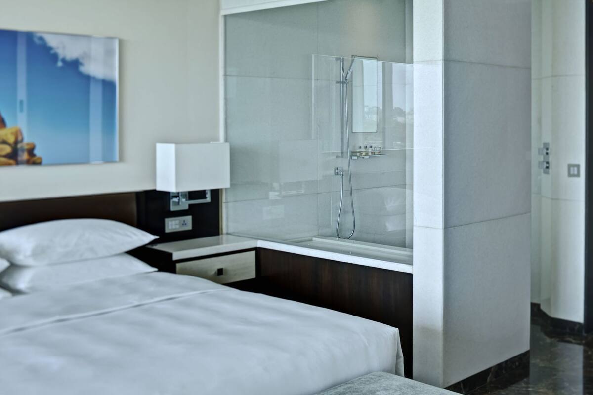 Une chambre d’hôtel moderne équipée d’un lit, d’une table de chevet, d’une lampe et d’une douche séparée par des parois en verre.
