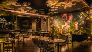 A restaurant with a mural of flowers and birds on the wall, wooden tables and chairs, and a bar area with shelves.