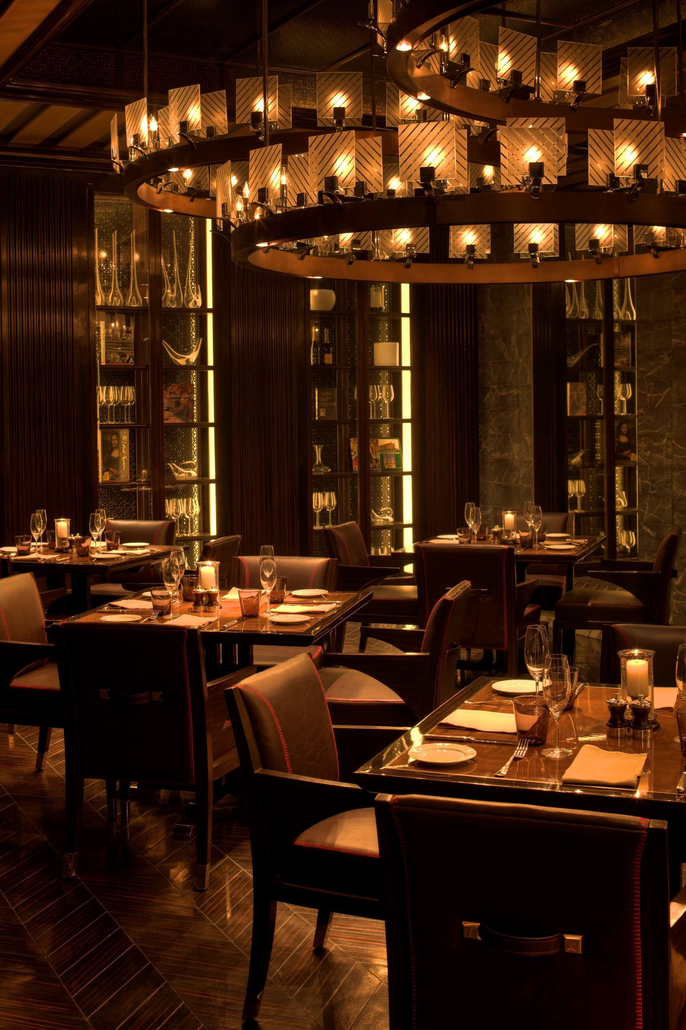 Un restaurante tenuemente iluminado con varias mesas y sillas, un candelabro central y estanterías con copas.