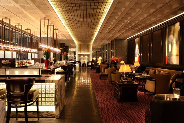 A spacious hotel lobby with a long bar, comfortable seating areas, and a patterned carpet.