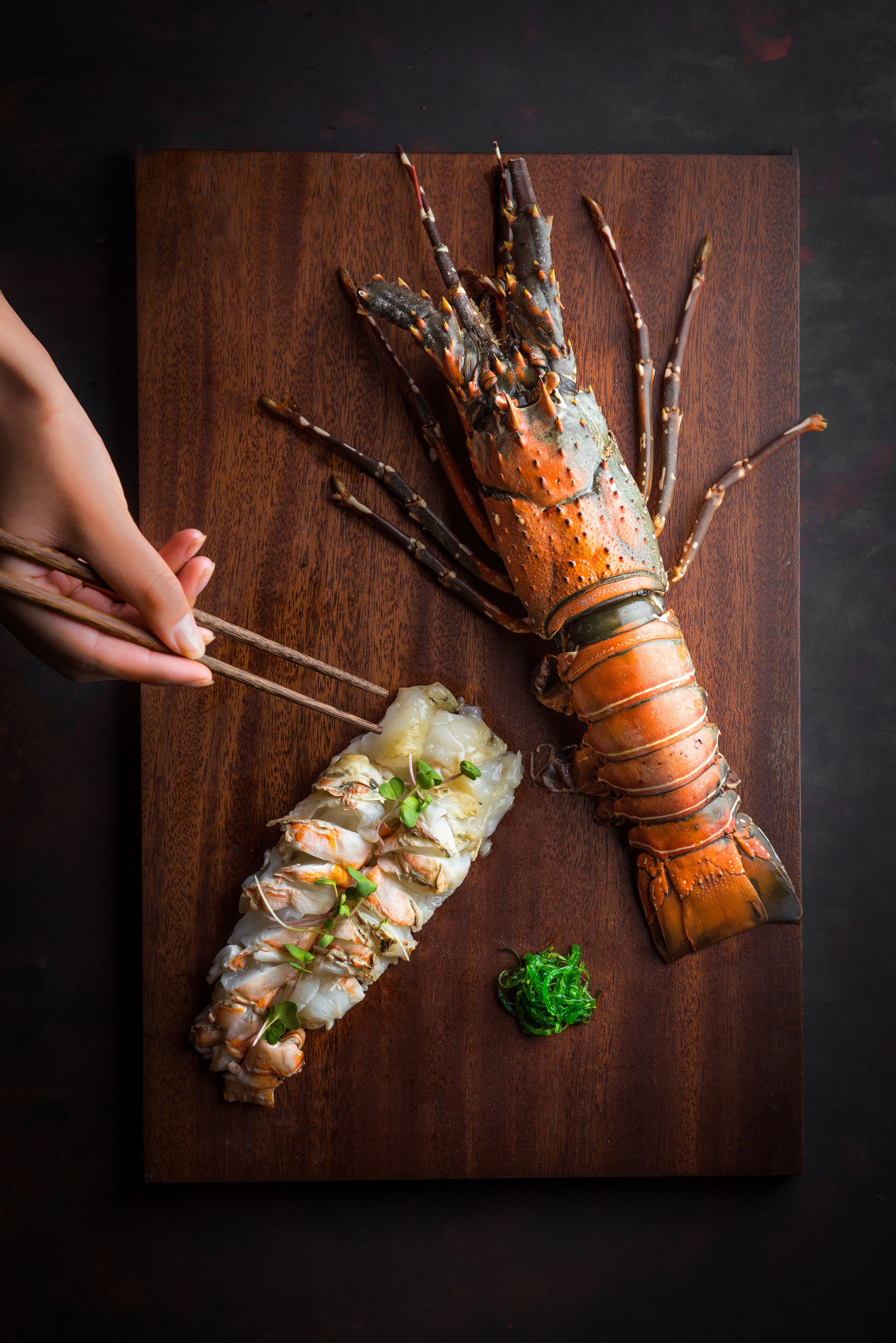 A lobster on a wooden board with chopsticks, a rolled lobster tail, and some green garnish.