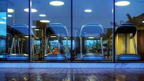 Un gimnasio con equipo de ejercicio visible a través de paredes de vidrio, suelo de madera y luces en el techo.