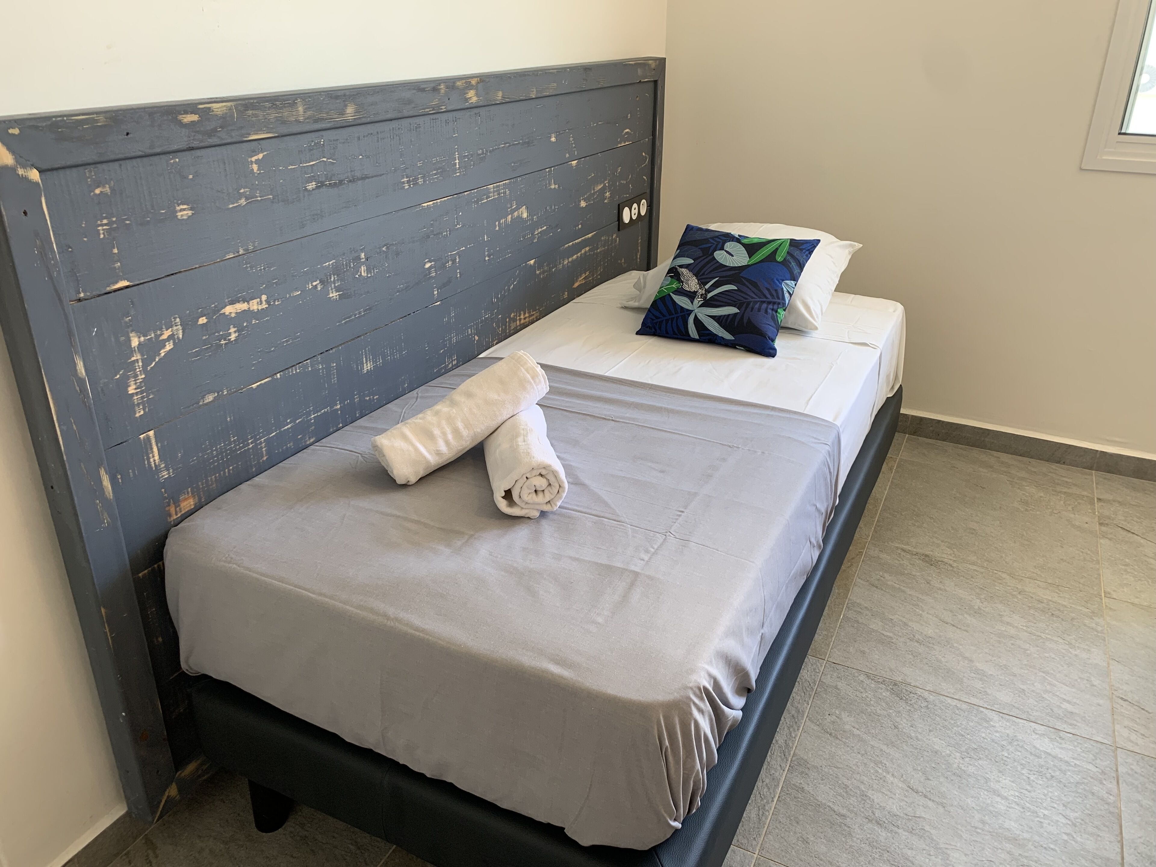 Una cama con una colcha gris, una toalla blanca enrollada y una almohada azul con un estampado de hojas.