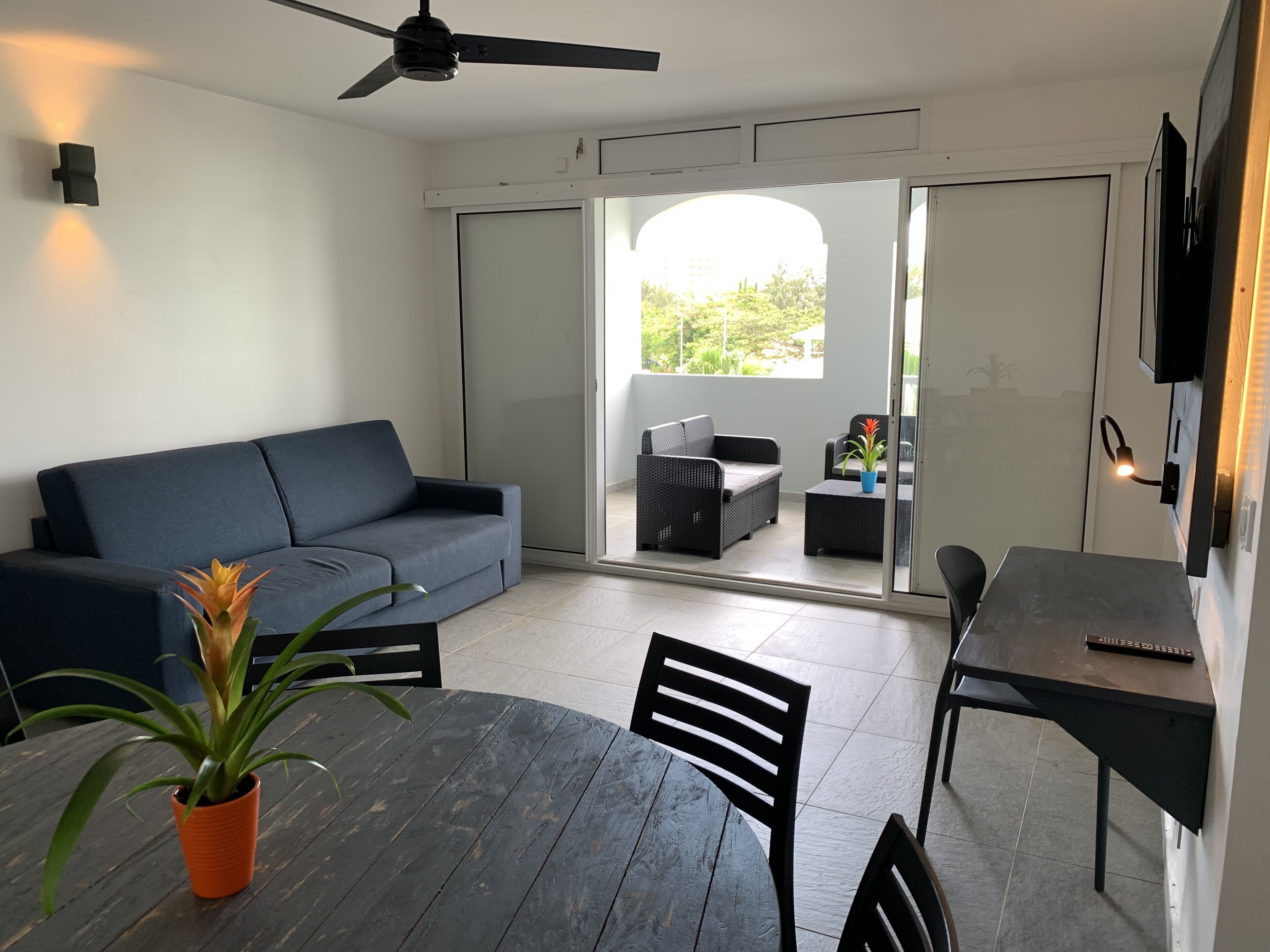 A modern living area with a dark blue sofa, a wooden dining table with chairs, a ceiling fan, and a view through glass doors to an outdoor patio with a wicker sofa and a potted plant.
