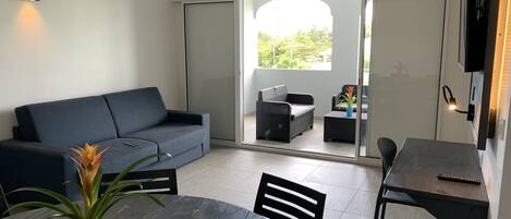 A modern living area with a dark blue sofa, a wooden dining table with chairs, a ceiling fan, and a view through glass doors to an outdoor patio with a wicker sofa and a potted plant.
