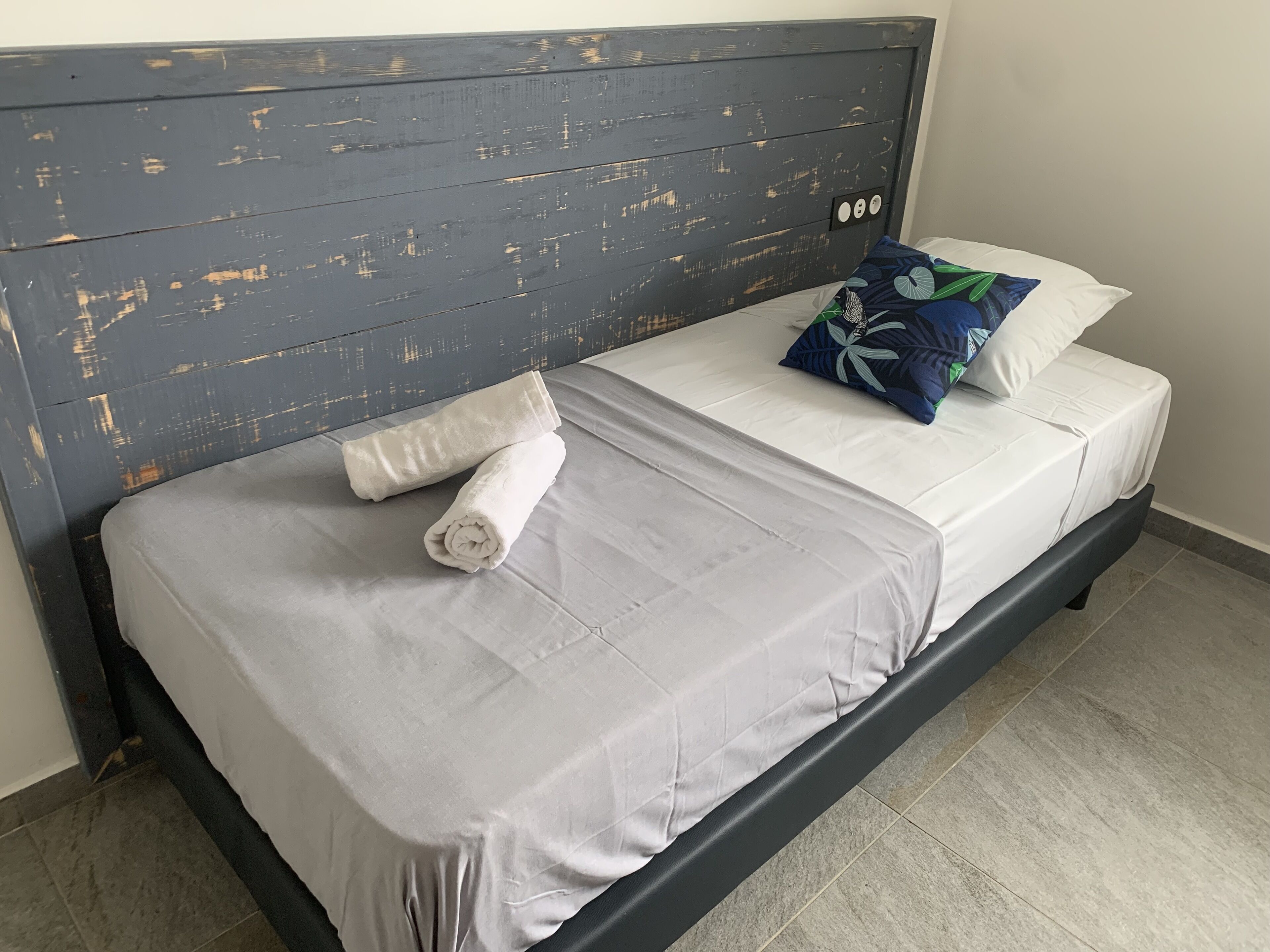 Un lit avec une couvre-lit grise, deux serviettes enroulées, un oreiller bleu orné d’un motif de feuilles et une tête de lit en bois au fini vieilli.