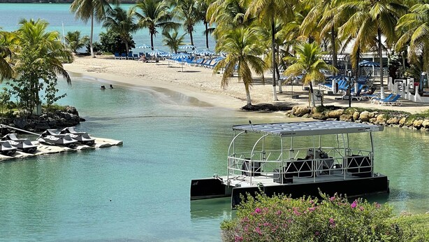 A beach with a boat docked near the shore, surrounded by palm trees and a sandy beach with lounge chairs and umbrellas. - Karaibes Residence (Le Gosier)