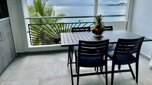 A balcony with a table and chairs, a fruit basket, and a view of the sea and palm trees. - Karaibes Residence (Le Gosier)