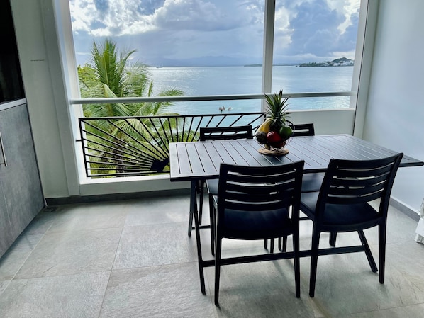 A balcony with a table and chairs, a fruit basket, and a view of the sea and palm trees.