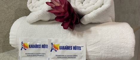 A bathroom with two white towels, a red flower, and two packages of Karāibes Hotel shampoo.