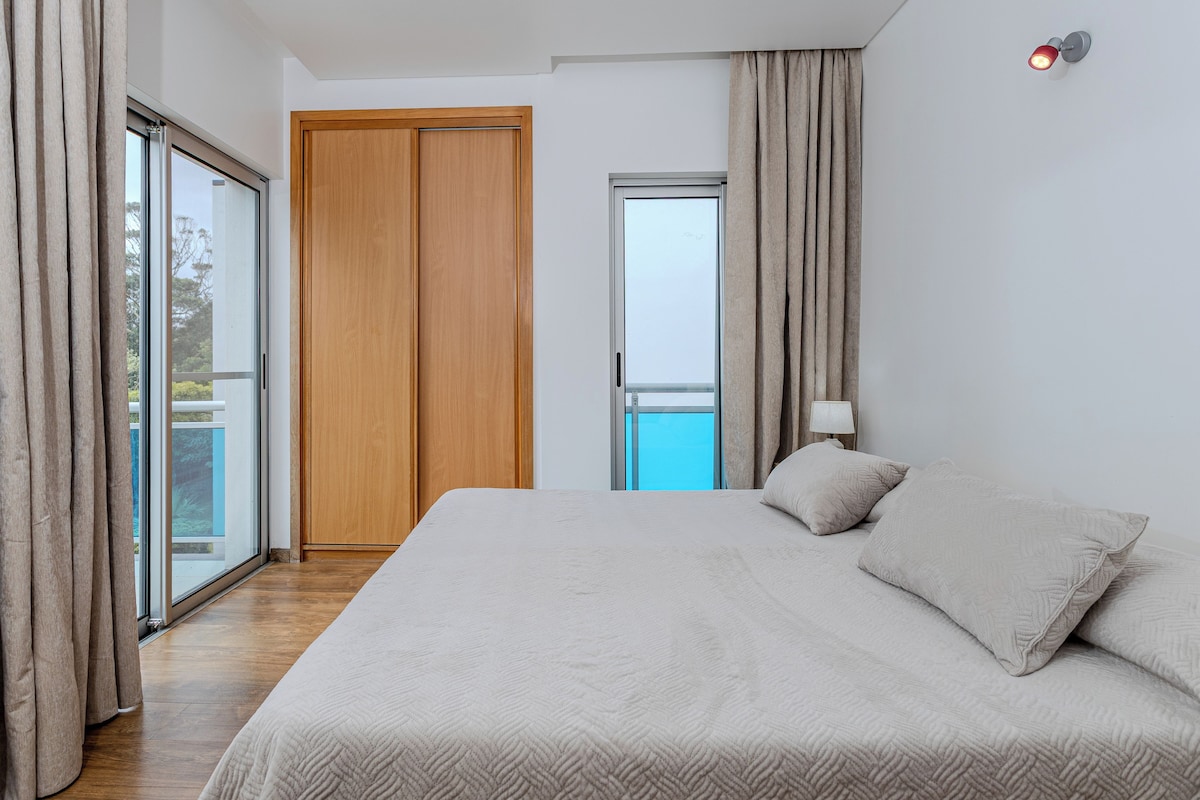 Un dormitorio con una cama grande, una puerta corrediza de vidrio, un armario de madera y una mesita de noche con lámpara.