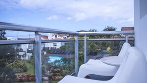 Ein Balkon mit weißen Stühlen, einer Glasreling und Blick auf einen Swimmingpool und einen Garten.