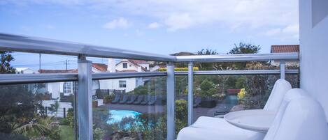 Un balcon avec des chaises blanches, une balustrade en verre et une vue sur une piscine et un jardin.